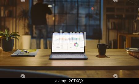 Shot of a Laptop on a Desk with Statistics Graph, Chart and Various Data, Showing Company Growth and Success. In the Background Businessman Drinks Stock Photo