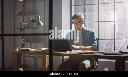 Creative Developer Works on a Laptop in His Studio. Designing Apps, Games and Doing Video Editing. In the Background Modern Sunny Loft Office with Stock Photo