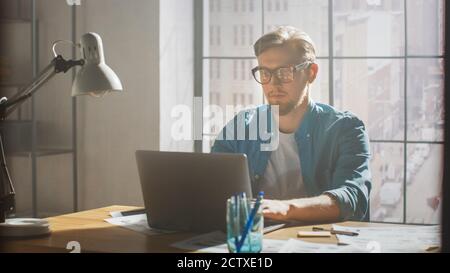 Creative Developer Works on a Laptop in His Studio. Designing Apps, Games and Doing Video Editing. In the Background Modern Sunny Loft Office with Stock Photo
