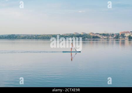 Young woman in a dress swims on the lake on surfboard Stock Photo