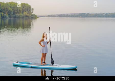 Young woman in a dress swims on the lake on surfboard Stock Photo
