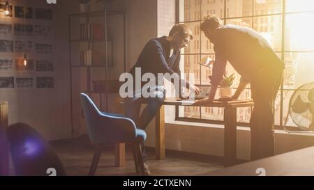 Two Young and Handsome Designers Sit on the Desk and Discuss Social Media App Interfaces on Their Laptops. They Work in a Sunny Creative Office Loft Stock Photo