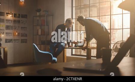 Two Young and Handsome Designers Sit on the Desk and Discuss Social Media App Interfaces on Their Laptops. They Work in a Sunny Creative Office Loft Stock Photo
