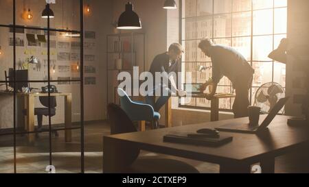 Two Young and Handsome Designers Sit on the Desk and Discuss Social Media App Interfaces on Their Laptops. They Work in a Sunny Creative Office Loft Stock Photo