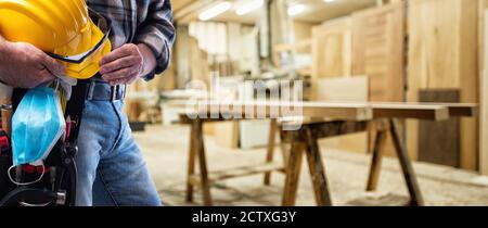 Carpenter in the carpentry workshop, holds helmet, goggles, leather gloves and surgical mask in hand to prevent Coronavirus infection. Preventing Pand Stock Photo