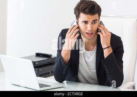 Overwork and job pressure. Stressed businessman speaking on two phones while working on a laptop at the office. Stock Photo