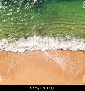 An aerial drone image looking down onto a sandy beach with white rolling waves and surf crashing onto a beach from an emerald green ocean in Cornwall, Stock Photo