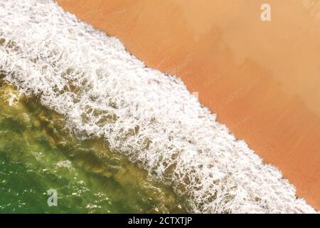 An aerial drone image looking down onto a sandy beach with white rolling waves and surf crashing onto a beach from an emerald green ocean in Cornwall, Stock Photo