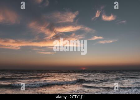 Sunset sun down hidden on horizon with stormy waves sea shore and epic colorful clouds lightened by warm sun rays Stock Photo