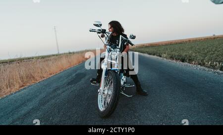 Shallow focus of a cool curly-haired male posing on his motorcycle on the road Stock Photo