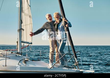Feeling happy together. Full length of a excited retired man and woman, senior couple in love standing on the side of yacht deck floating in sea Stock Photo