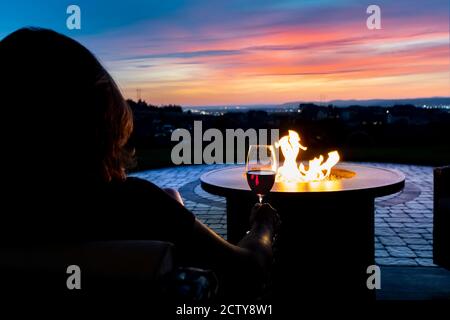 A woman relaxes with a glass of wine at sunset by a fire pit on the patio of a luxury home overlooking the Spokane valley and city in Spokane, Wa USA Stock Photo