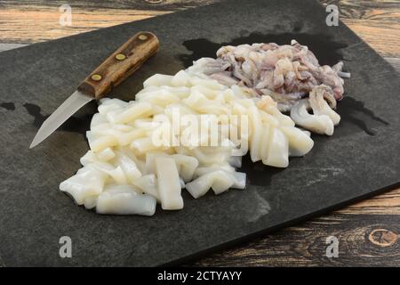 Preparing and cutting raw squid tubes and tentacles on weathered black cutting board with kitchen knife Stock Photo