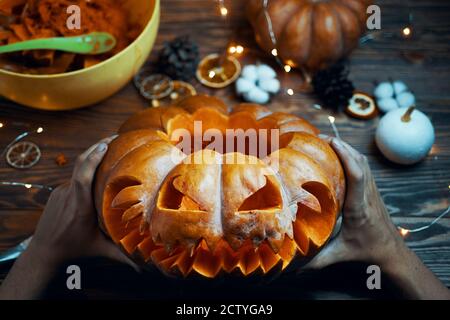 Autumn composition of witch holding Halloween pumpkin. The concept of Halloween with the carving of faces on the pumpkin. Bowl for seeds and waste. Garland, dried orange slices on wooden background. Stock Photo