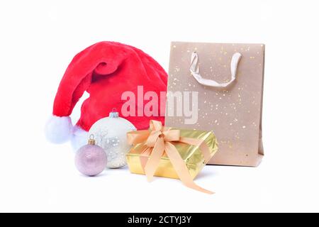 Close up of a gift box surrounded by Christmas baubles, a Santa hat and a gift paper bag on a light background. Christmas holiday and gifts concept. Stock Photo