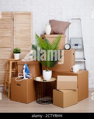 moving day concept - stacked cardboard boxes, houseplant and other domestic things over white brick wall background Stock Photo