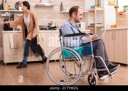Invalid man in wheelchair working remotely on laptop in kitchen while is standing in front of refrigerator. Disabled paralyzed handicapped man with walking disability integrating after an accident. Stock Photo