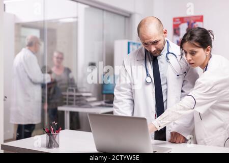 Team of young doctors working together in hospital checking patient evolution on laptop. Elderly aged woman and senior medic on clinic corridor. Physician wearing white coat and stethoscope. Stock Photo