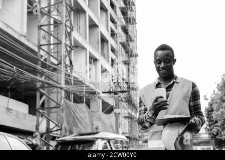 Happy young African man construction worker using phone while holding clipboard at building site Stock Photo