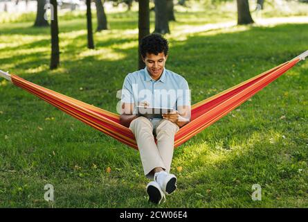 Young smiling man sitting on a hammock in park and drawing on digital tablet Stock Photo