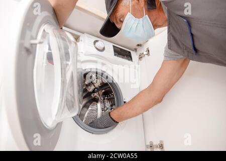 Working man in medical mask plumber repairs washing machine in kitchen Stock Photo