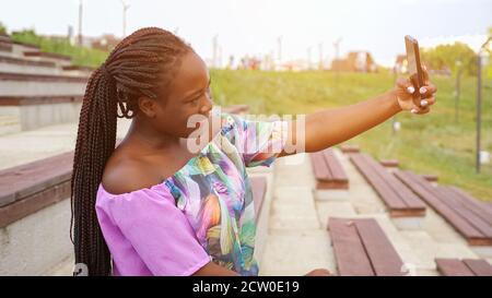 attractive young African-American lady with long ponytail in stylish color blouse makes selfie with smartphone sitting on bench in green summer park Stock Photo