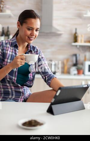 Enjoying cup of green tea while using tablet pc in kitchen. Working from home using device with internet technology, typing, on gadget during breakfast. Stock Photo
