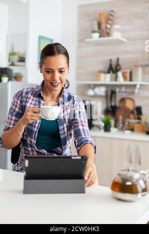 Woman smiling using tablet pc in the morning and enjoying a cup of hot green tea. Working from home using device with internet technology, typing, on gadget during breakfast. Stock Photo