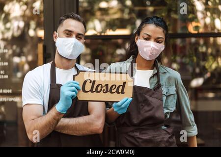 Man and woman wearing medical face masks holding closed sign while standing in front of a cafe Stock Photo