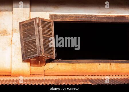 Broken wooden shutter in cracked white concrete wall of an old abandoned house, ancient town in Champasak, South Laos. Stock Photo