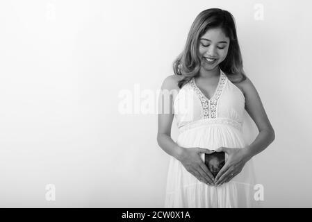 Studio shot of young happy Asian pregnant woman smiling and looking at sonogram of her baby on her stomach Stock Photo