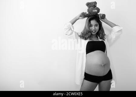 Studio shot of young happy Asian pregnant woman holding teddy bear with heart and love sign on top on her head Stock Photo