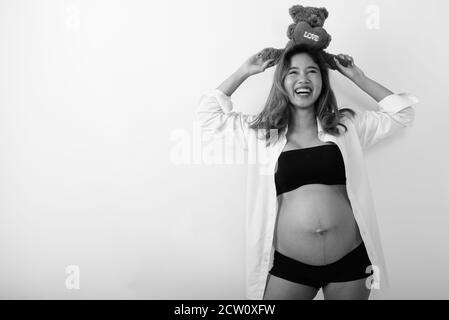 Happy young Asian pregnant woman thinking while holding teddy bear with heart and love sign on top on her head Stock Photo