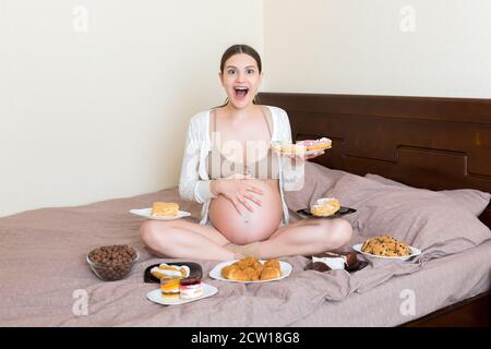 Hungry pregnant woman sitting on the bed is eating a lot of unhealthy food such as croissants, donuts, chocolate cereal balls, cakes and cookies. Swee Stock Photo