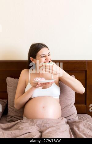 Pregnant woman is eating many donuts relaxing in bed. Unhealthy dieting during pregnancy concept. Stock Photo