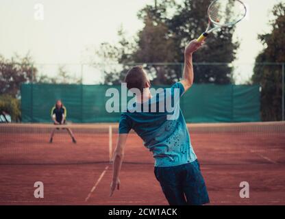 Rear view of a man wearing a blue shirt and blue pants jumping and swining his racket while serving in a match of tennis. Playing outside of an orange Stock Photo