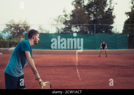 Back view of a man holding a racket and a ball, prepairing to serve while playing a match of tennis outside on an orange clay field Stock Photo