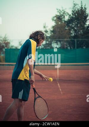 Split Croatia September 2020 Back view of a man holding a racket and a ball, prepairing to serve while playing a match of tennis outside on an orange Stock Photo