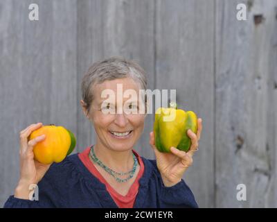 Modern middle-aged Caucasian Canadian country woman with short hair holds two large organic bell peppers in her hands. Stock Photo
