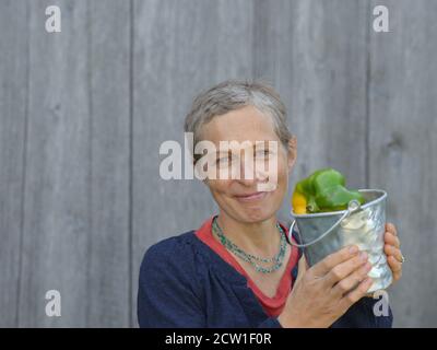 Modern middle-aged Caucasian Canadian country woman with short hair shows a small bucket full of homegrown large organic bell peppers.