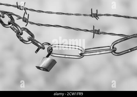 Padlock and steel chains with barbed wire against clouded sky symbolizing lockdown in monochrome Stock Photo