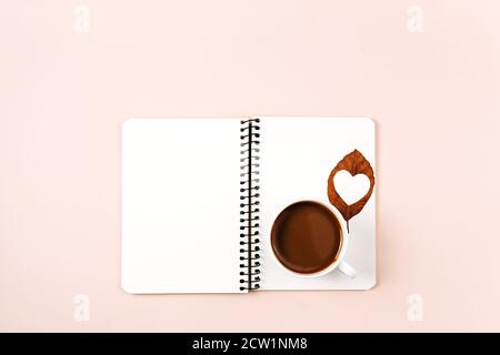 Flat lay composition. Notebook, cup coffee, dried leaves with heart carved in middle on pink background for advertisement. Creative autumn, thanksgivi Stock Photo