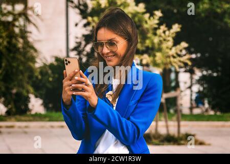 Smiling businesswoman in blue blazer holding smartphone. Typing message. Texting Stock Photo