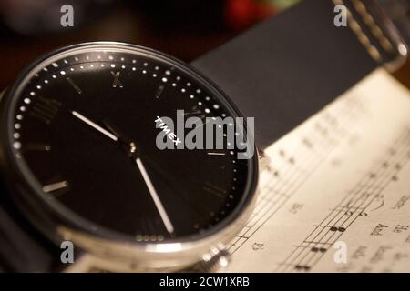 Timex analog wrist watch laying on sheet music. Black leather strap with silver roman numerals. Stock Photo