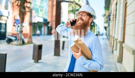 Young redhead architect man with long beard wearing hardhat smiling happy. Having conversation talking on the smartphone holding blueprints at street Stock Photo