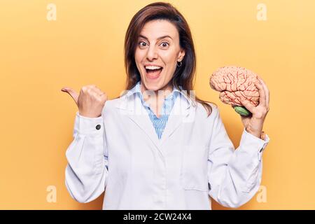 Young beautiful woman wearing doctor coat holding brain pointing thumb up to the side smiling happy with open mouth Stock Photo