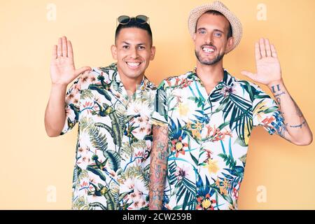 Young gay couple of two men wearing summer hat and hawaiian shirt waiving saying hello happy and smiling, friendly welcome gesture Stock Photo