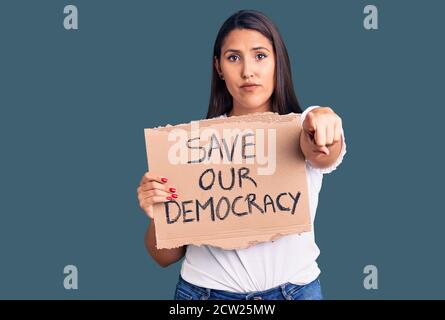 Young beautiful woman holding save our democracy cardboard banner pointing with finger to the camera and to you, confident gesture looking serious Stock Photo