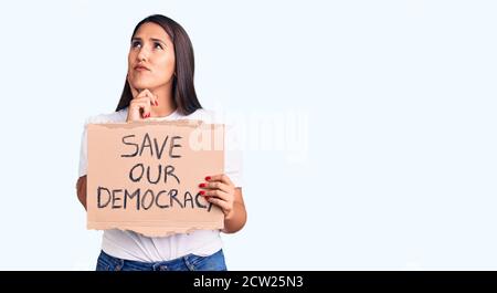 Young beautiful woman holding save our democracy cardboard banner serious face thinking about question with hand on chin, thoughtful about confusing i Stock Photo