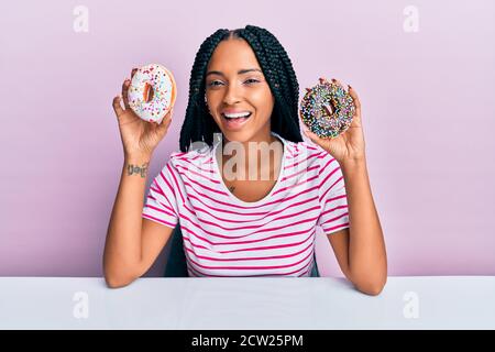 Beautiful hispanic woman holding tasty colorful doughnuts smiling and laughing hard out loud because funny crazy joke. Stock Photo